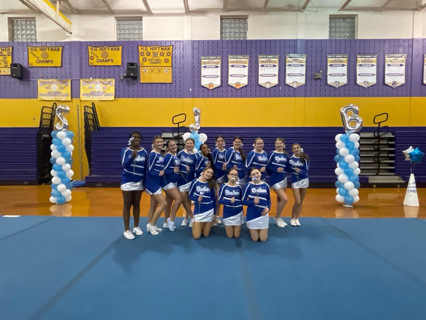 Cheerleaders in blue and white uniforms pose on a blue mat in a gym with 
