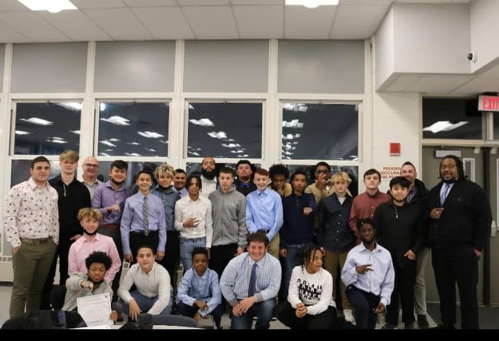 Group of people standing indoors, posing for a photo.  A few adults surround a larger group of teenagers, with large windows behind them.