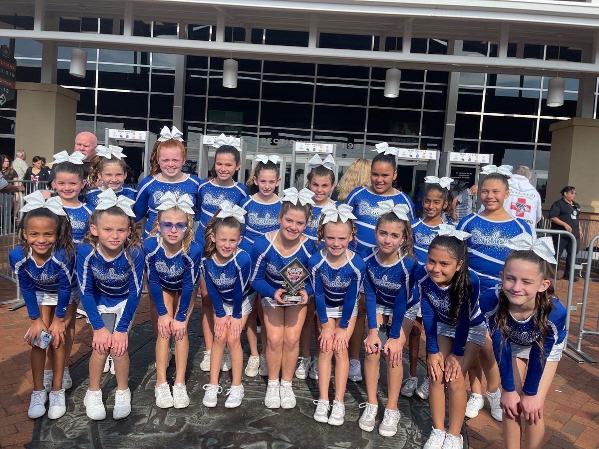 Cheerleading team in blue and white uniforms holding a trophy outdoors, smiling near a building.