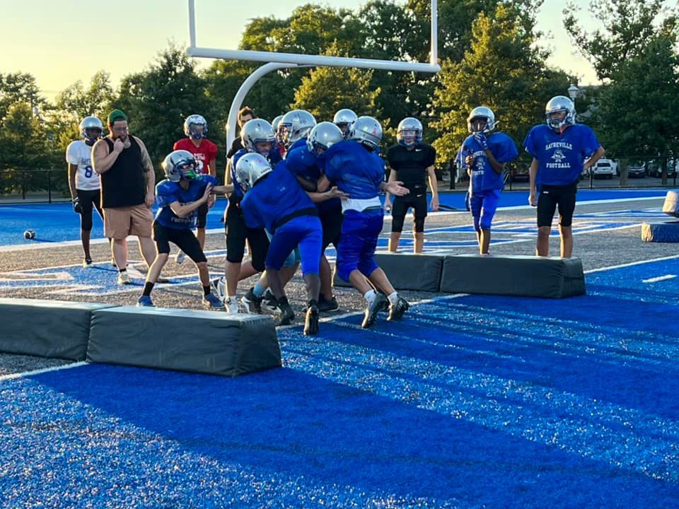 Football team practicing tackling on a blue turf field. Players in blue uniforms, coach watching.