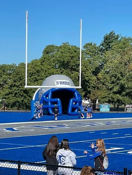 Football field with goalpost and blue helmet-shaped tunnel. People watch from the sidelines.
