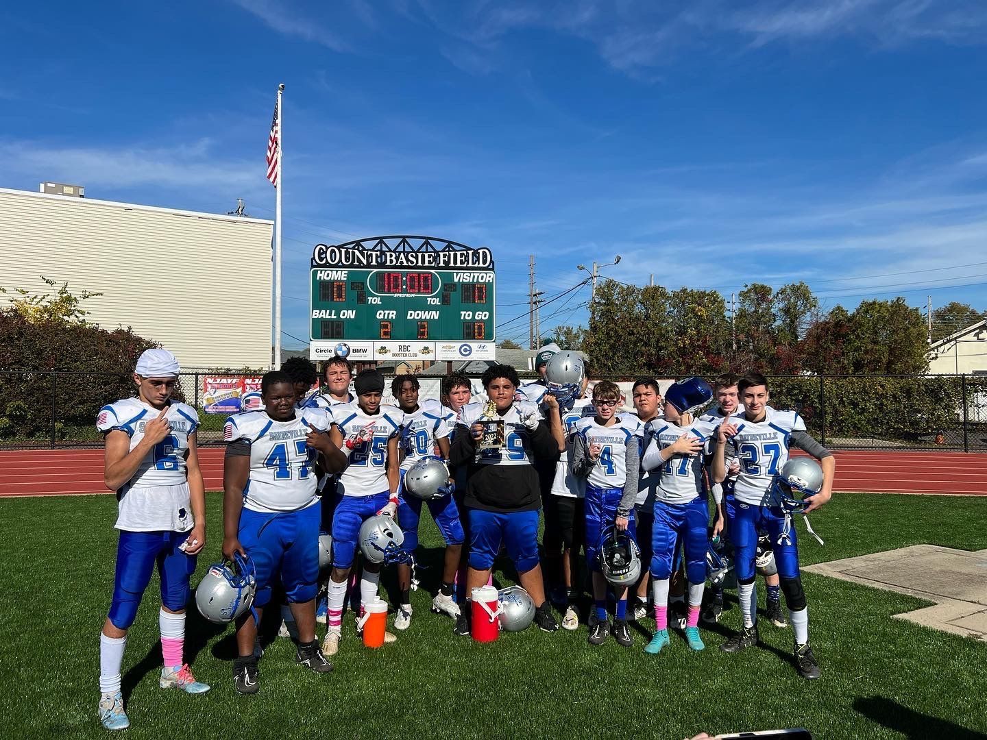 Youth football team in blue and white uniforms pose on a field with a scoreboard under a blue sky.