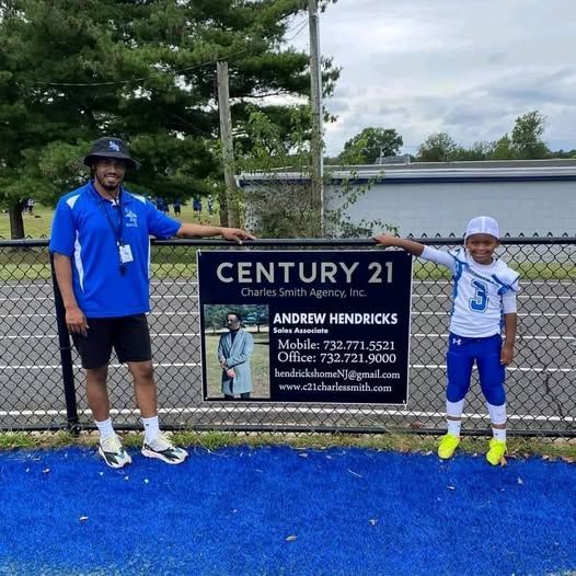 Man and child in a football uniform stand by a Century 21 sign on a blue turf field.