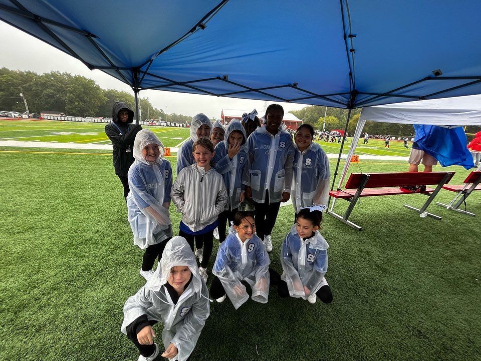 Group of children in raincoats under a blue tent on a green field. It's raining.