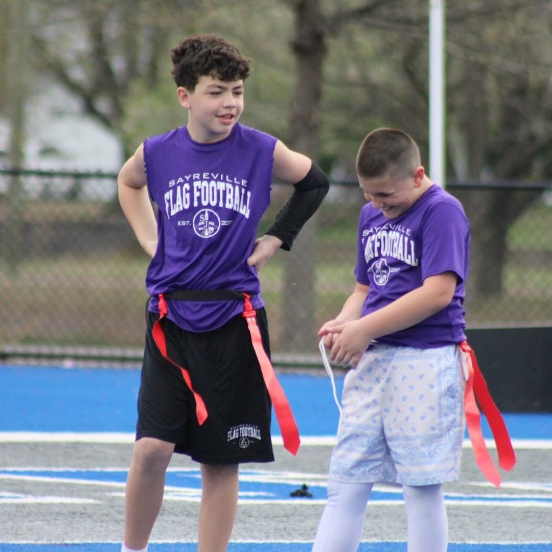 Two boys in purple flag football jerseys on a blue field. One has his hands on his hips, the other is holding a flag.