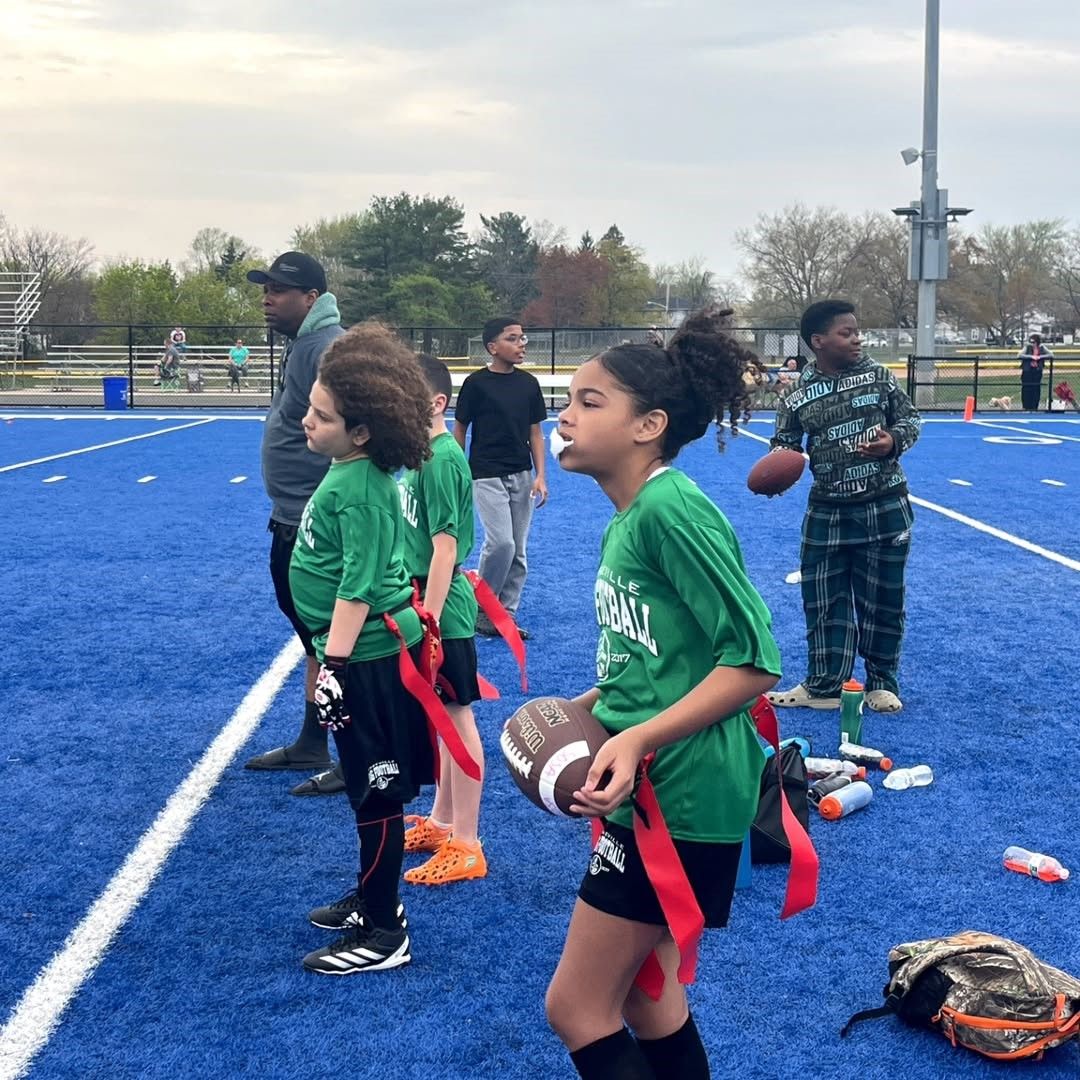 Children in green shirts with flags on a blue field, holding footballs, with a coach nearby.