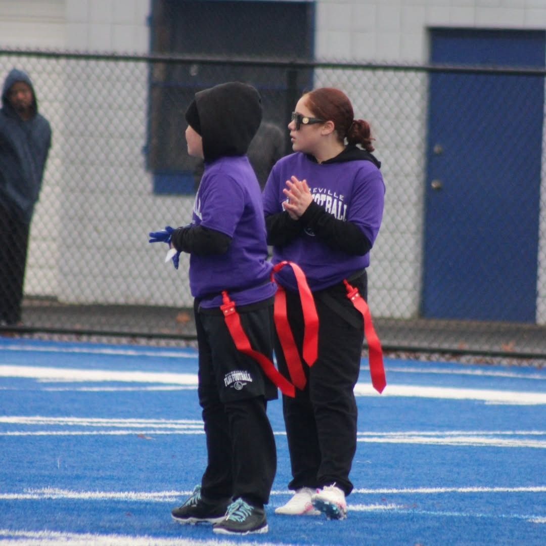 Two people in purple shirts and black pants stand on a blue field, holding red flags, with one applauding.