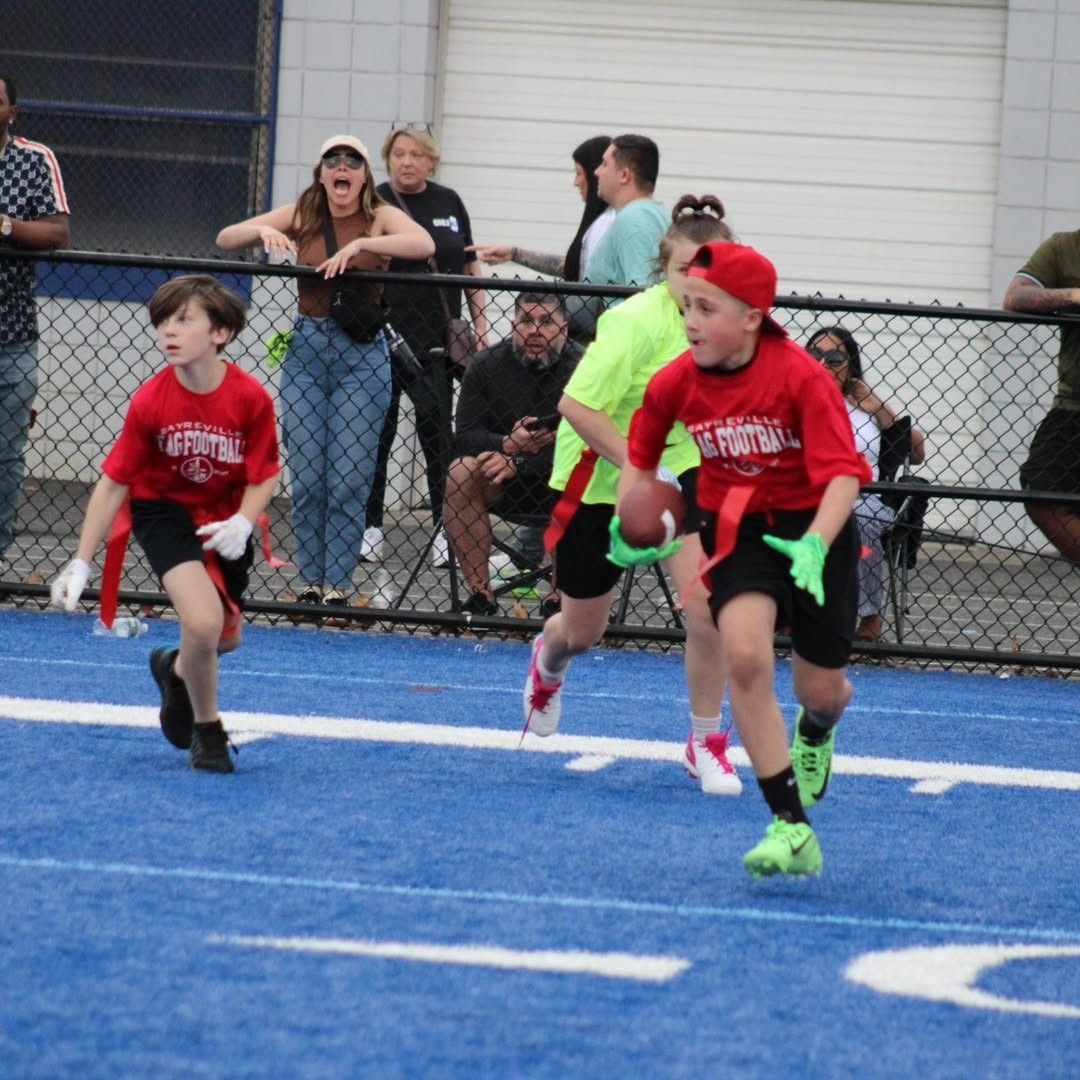 Two young players in red jerseys run with a football on a blue field during a flag football game.