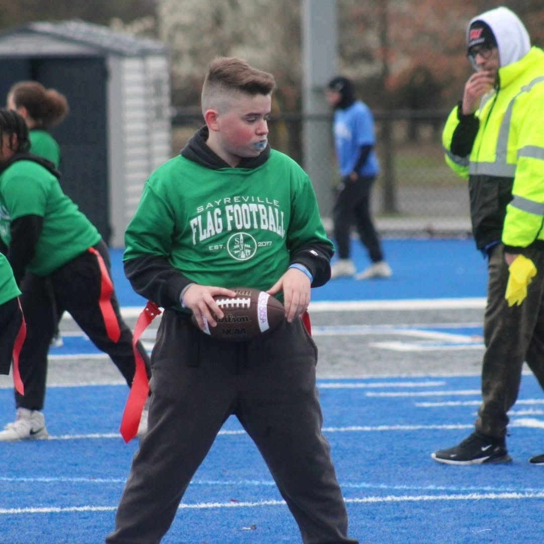 A boy in a green jersey holding a football on a blue field during a flag football game.