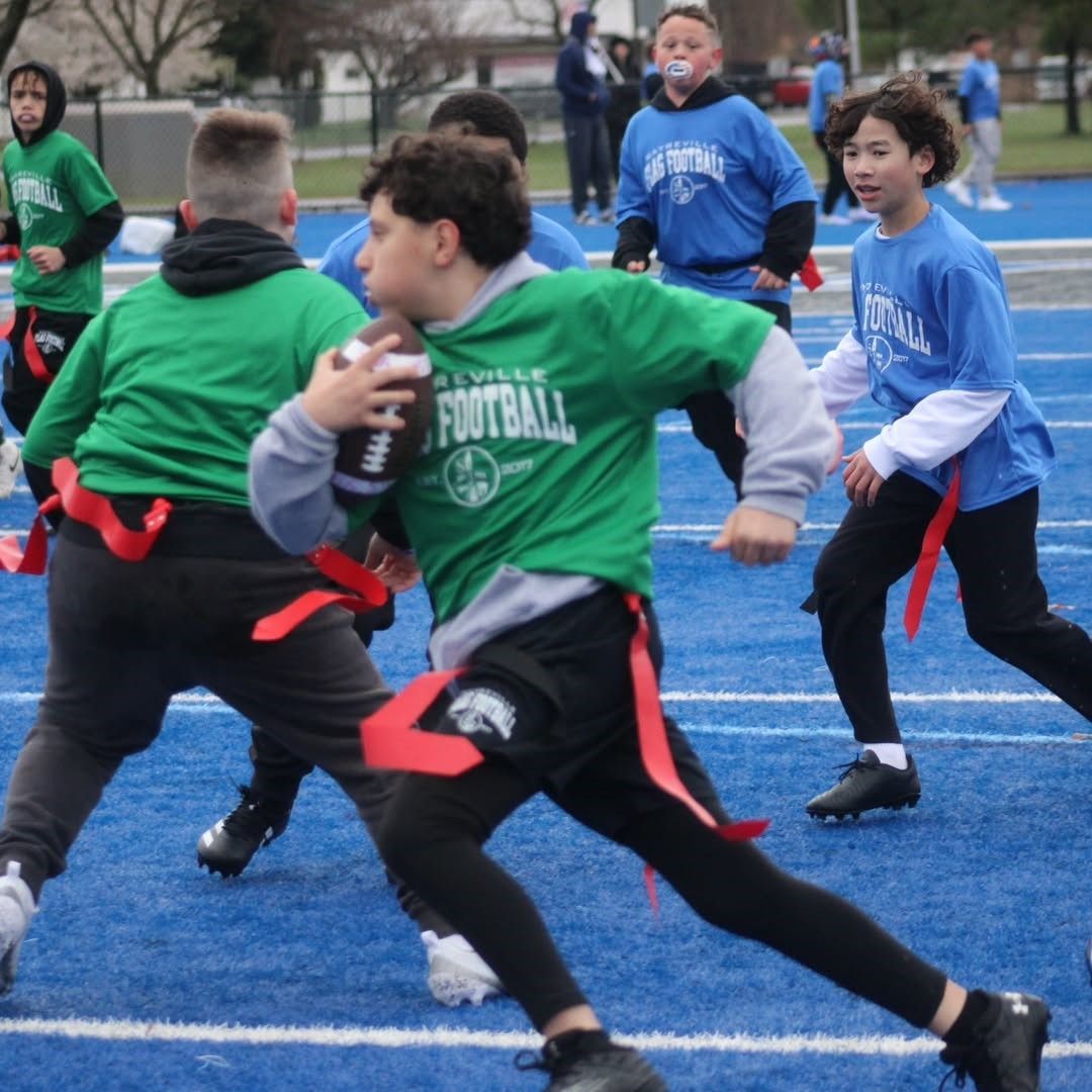 Children playing flag football on a blue field; a player in green shirt runs with the ball.