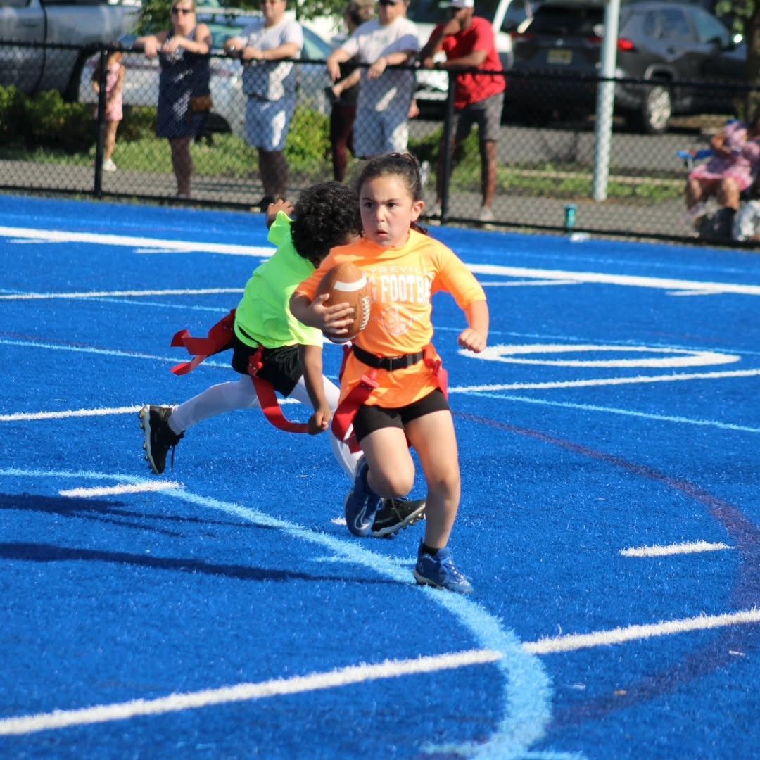 Girl in orange jersey runs with football pursued by another player on blue field.