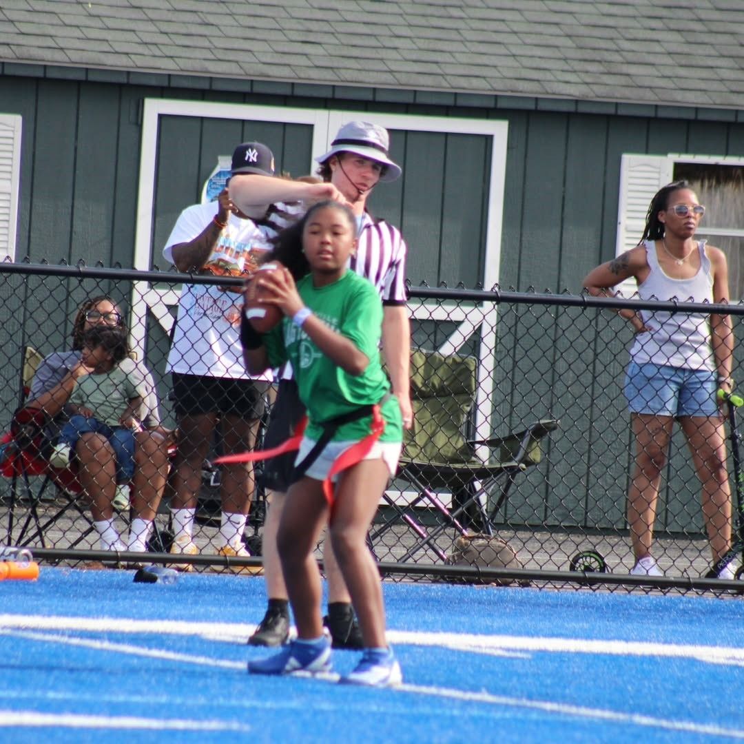 Young person in green jersey throws football on blue field, game in progress, referee nearby.