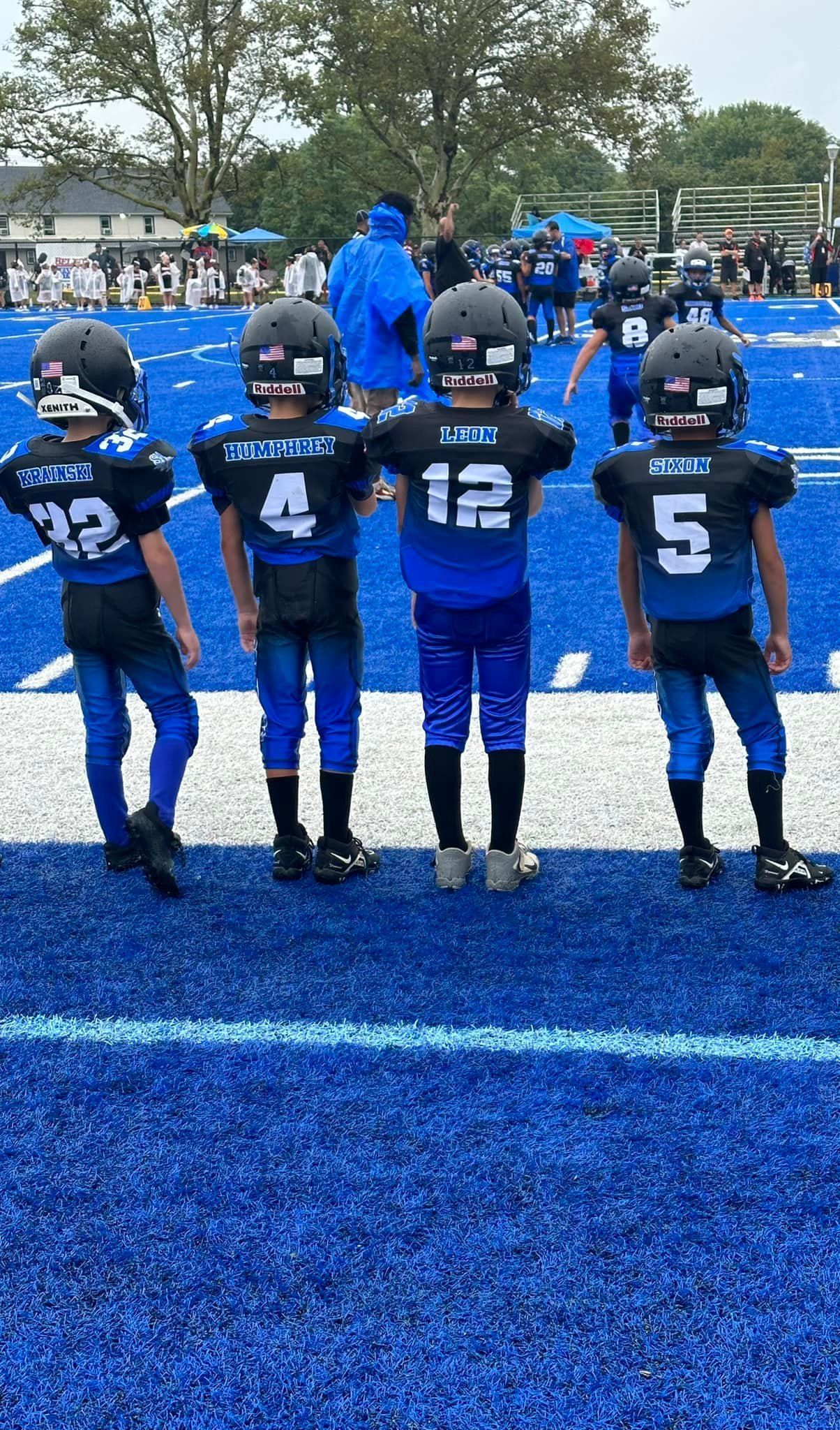 Four young football players in blue and black uniforms stand on a blue field.
