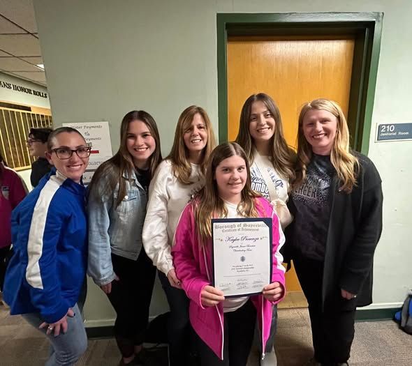 Group of people posing with an award in a hallway; the person holding it wears a pink jacket.