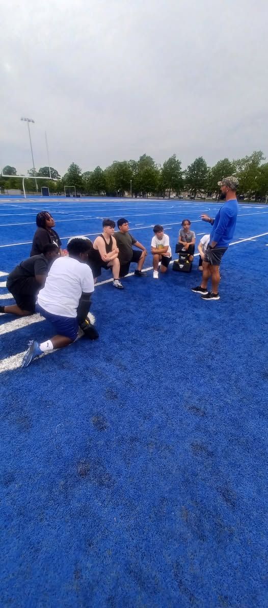 A group of people kneel on a blue track field while a man stands and gestures. Overcast sky.
