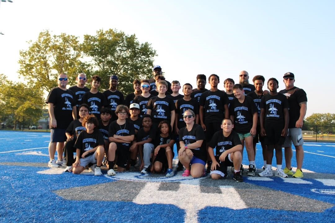 Group of people in black shirts posing on a blue field with large white markings, under sunlight.