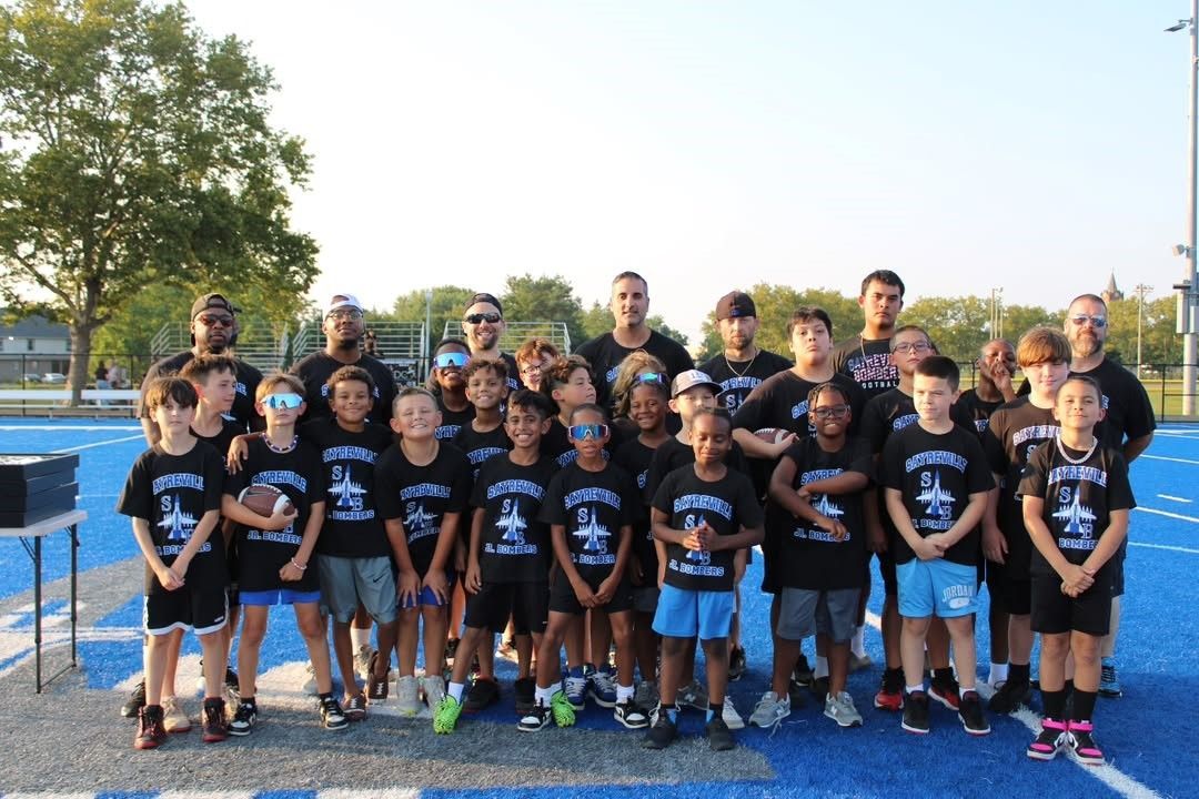 Football team, in black shirts, poses on a blue field. Some wear sunglasses and hats.