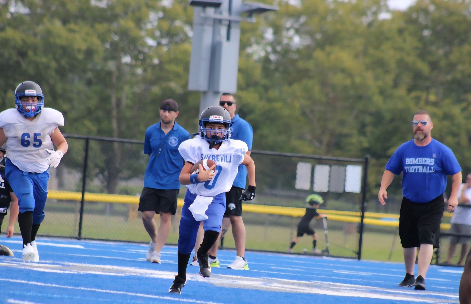 Football player running with ball on a blue field; teammates and coaches in background.