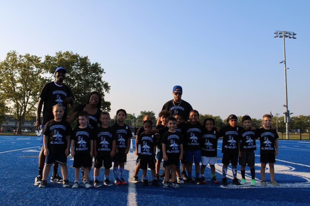 Youth football team posing on a blue field. Players and coaches wear matching black shirts and shorts.