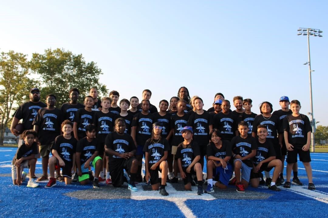 Group of people in black t-shirts on a blue field; a few kneeling. T-shirts have a plane logo.
