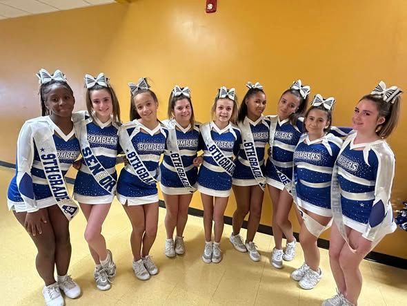 Cheerleaders in blue and white uniforms pose in a room. Some wear sashes that say 