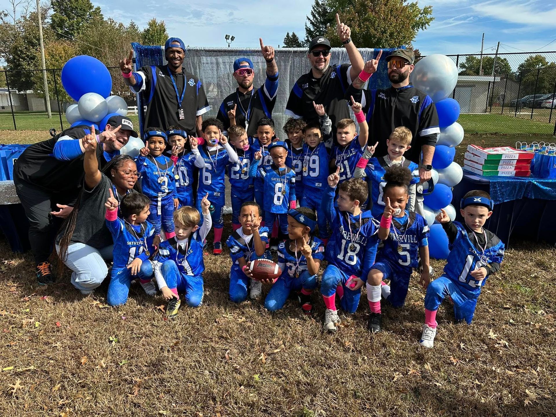 Youth football team in blue uniforms poses with coaches outdoors, celebrating. Blue and white balloons, backdrop.