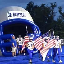 Football players entering a blue inflatable helmet tunnel, holding flags. 