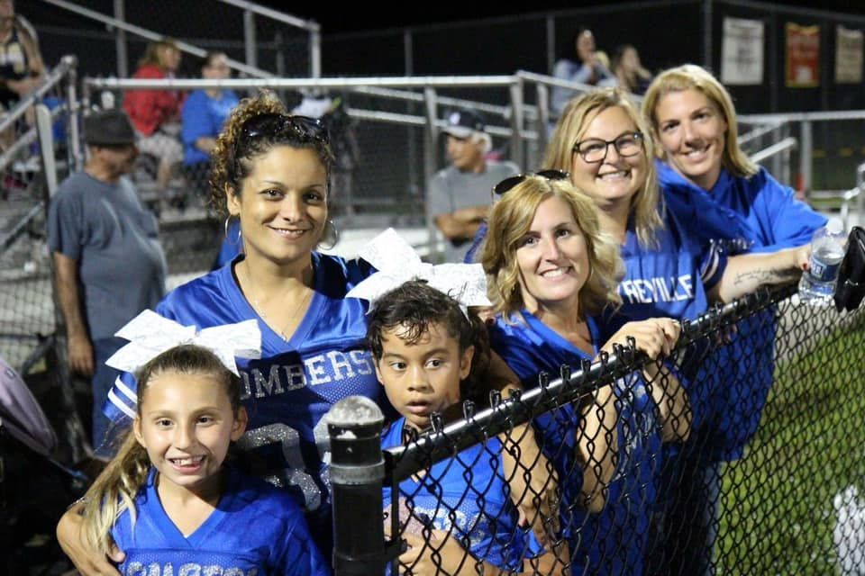 Fans in blue jerseys and cheerleading bows at a football game, leaning on a fence.