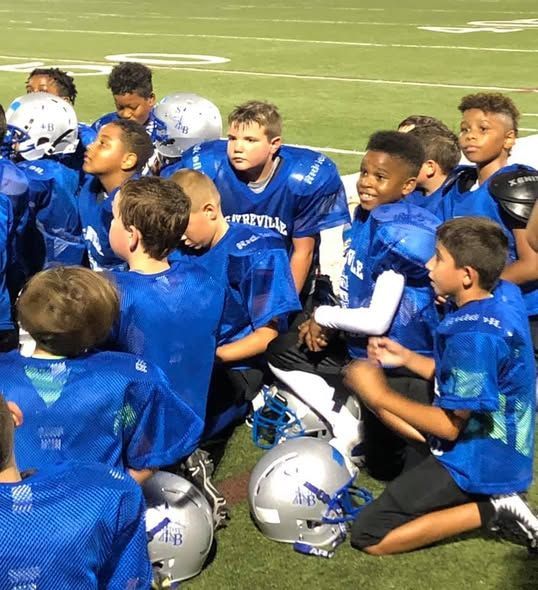 Youth football team in blue jerseys huddled together on the field, helmets nearby.