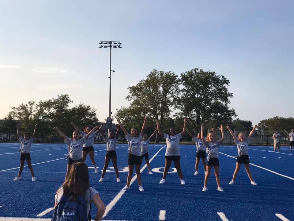 Cheerleaders doing jumping jacks on a blue football field with overhead lights.
