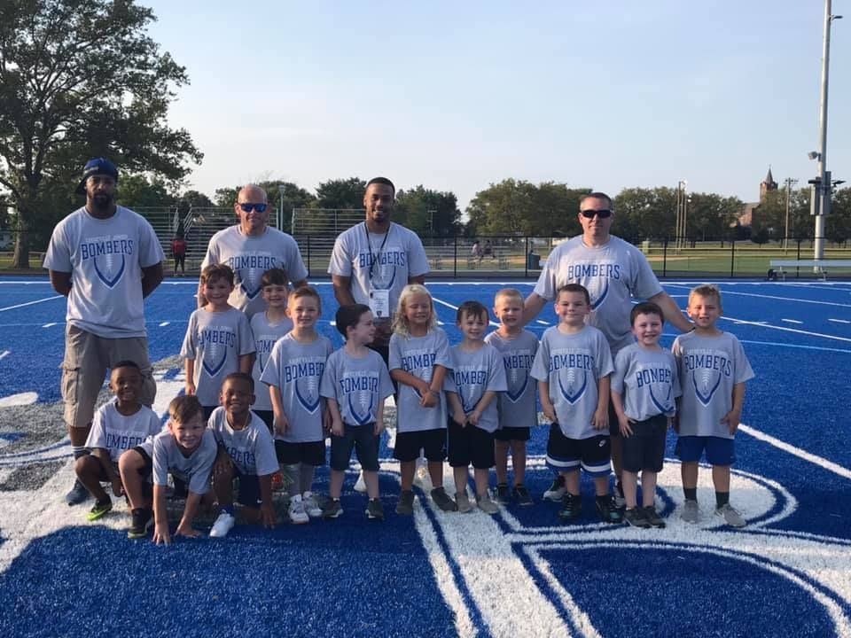 Youth football team and coaches pose on a blue field in matching gray shirts with team logo.