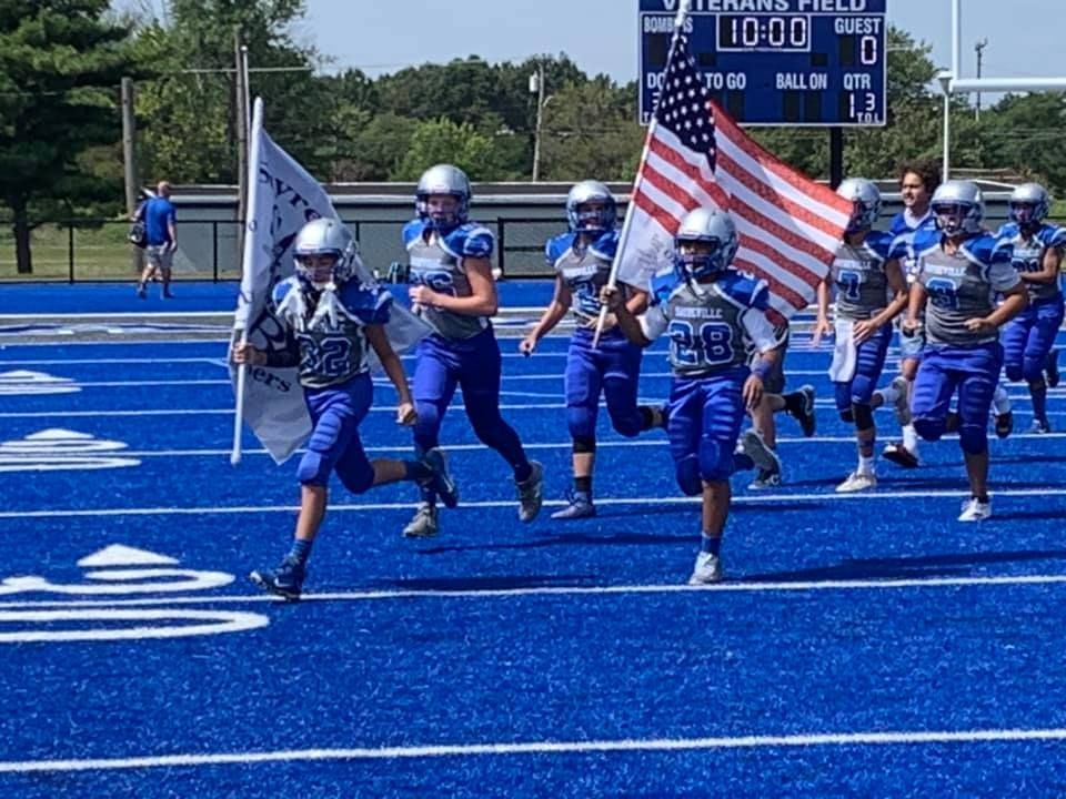 Football team runs onto a blue field, carrying flags, including an American flag.