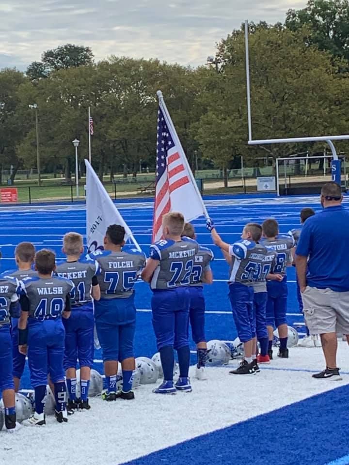 Youth football team on a blue field holding American and POW/MIA flags.