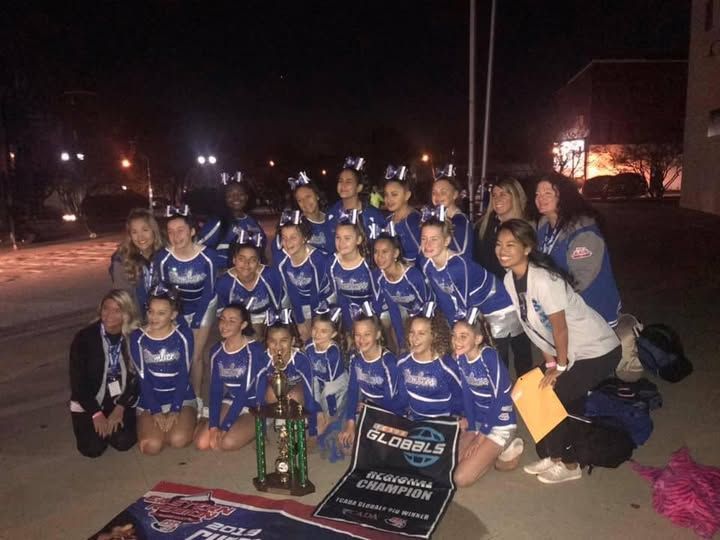 Cheerleading team in blue and white uniforms celebrating a competition win, holding trophy and banner at night.