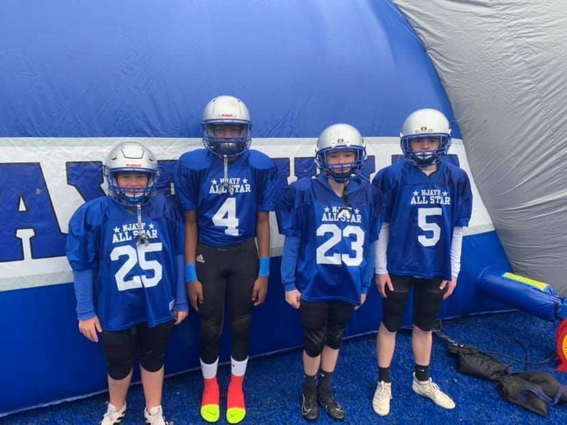 Four youth football players in blue jerseys and helmets pose in front of a blue inflatable tunnel.