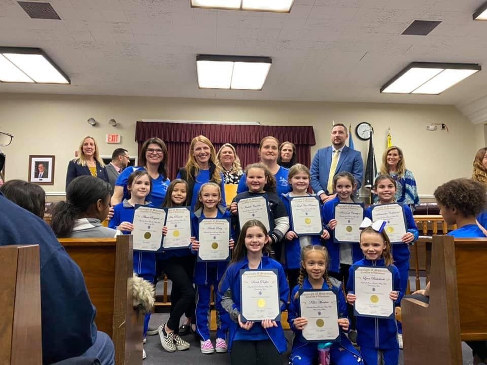 Group of children in blue uniforms holding certificates, posed with adults in a room.