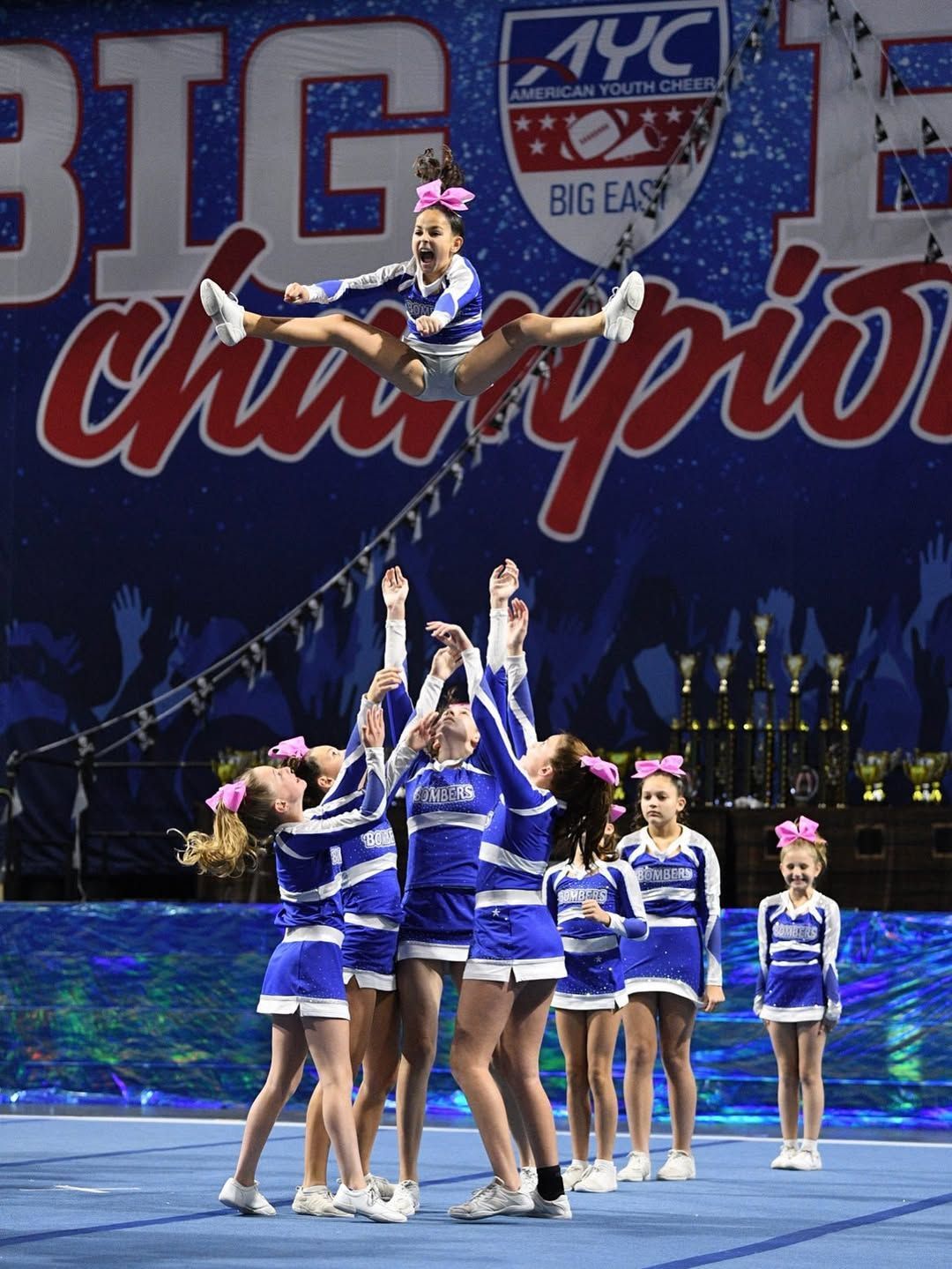 Cheerleaders in blue uniforms performing a stunt; one flyer jumps in the air above a pyramid, indoors.