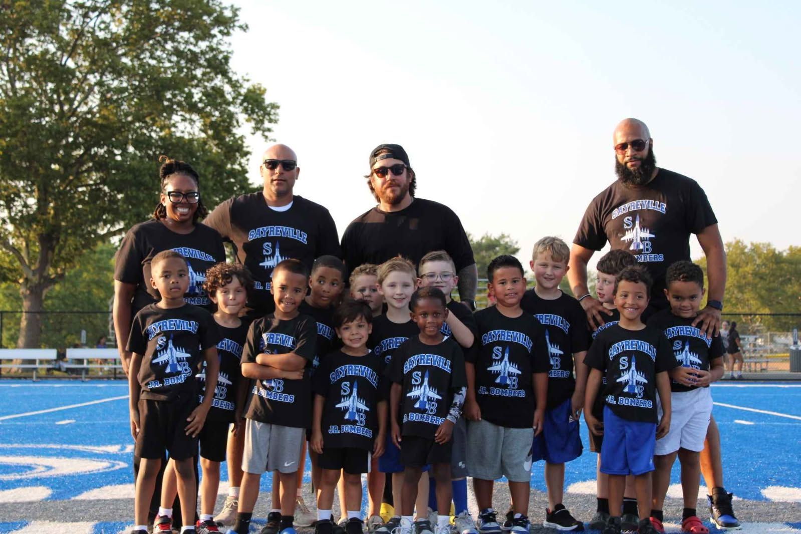 Youth football team poses with coaches on a blue track, all wearing matching black shirts with a jet graphic.