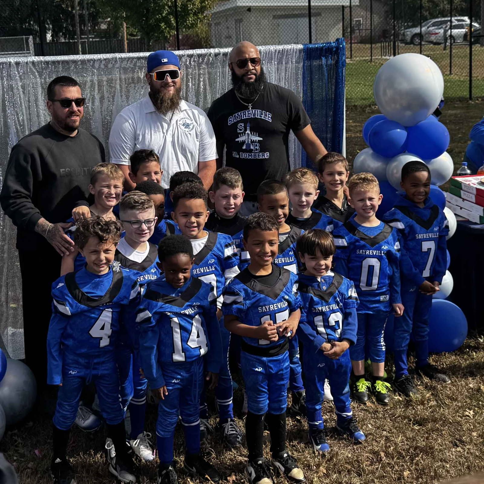 Football team posing with coaches at outdoor event; blue and silver theme.