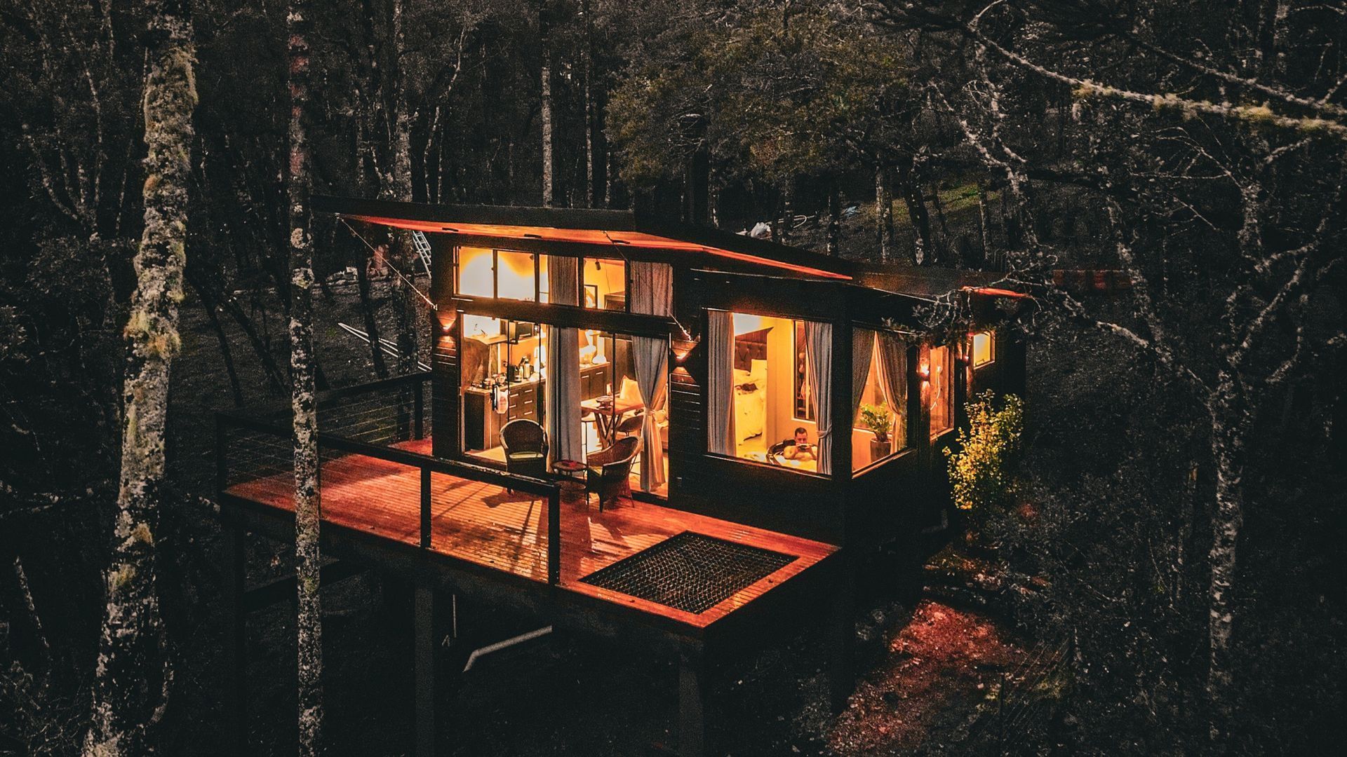 Cabana aconchegante em uma floresta escura ao entardecer, com luzes quentes iluminando o interior e o deck.