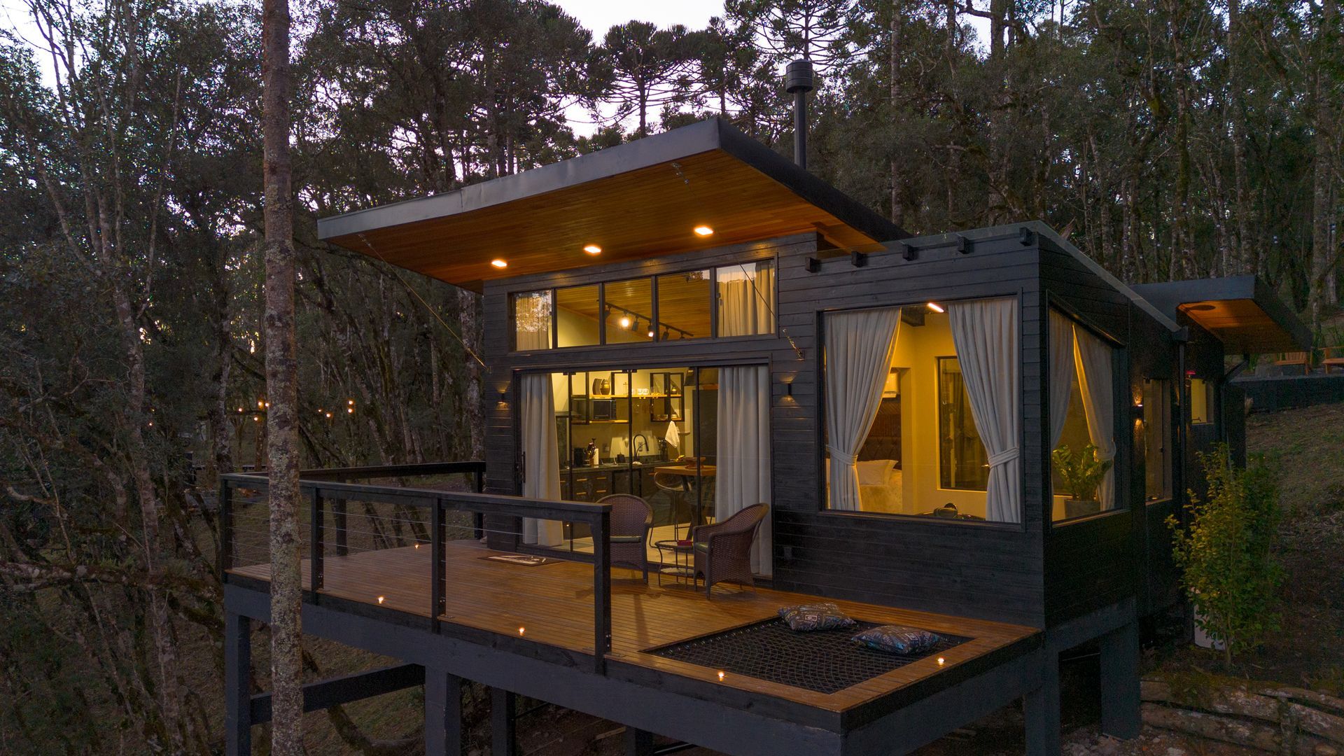 Cabana moderna com deck, aninhada entre as árvores. Exterior cinza, janelas amplas, iluminação interna aconchegante.