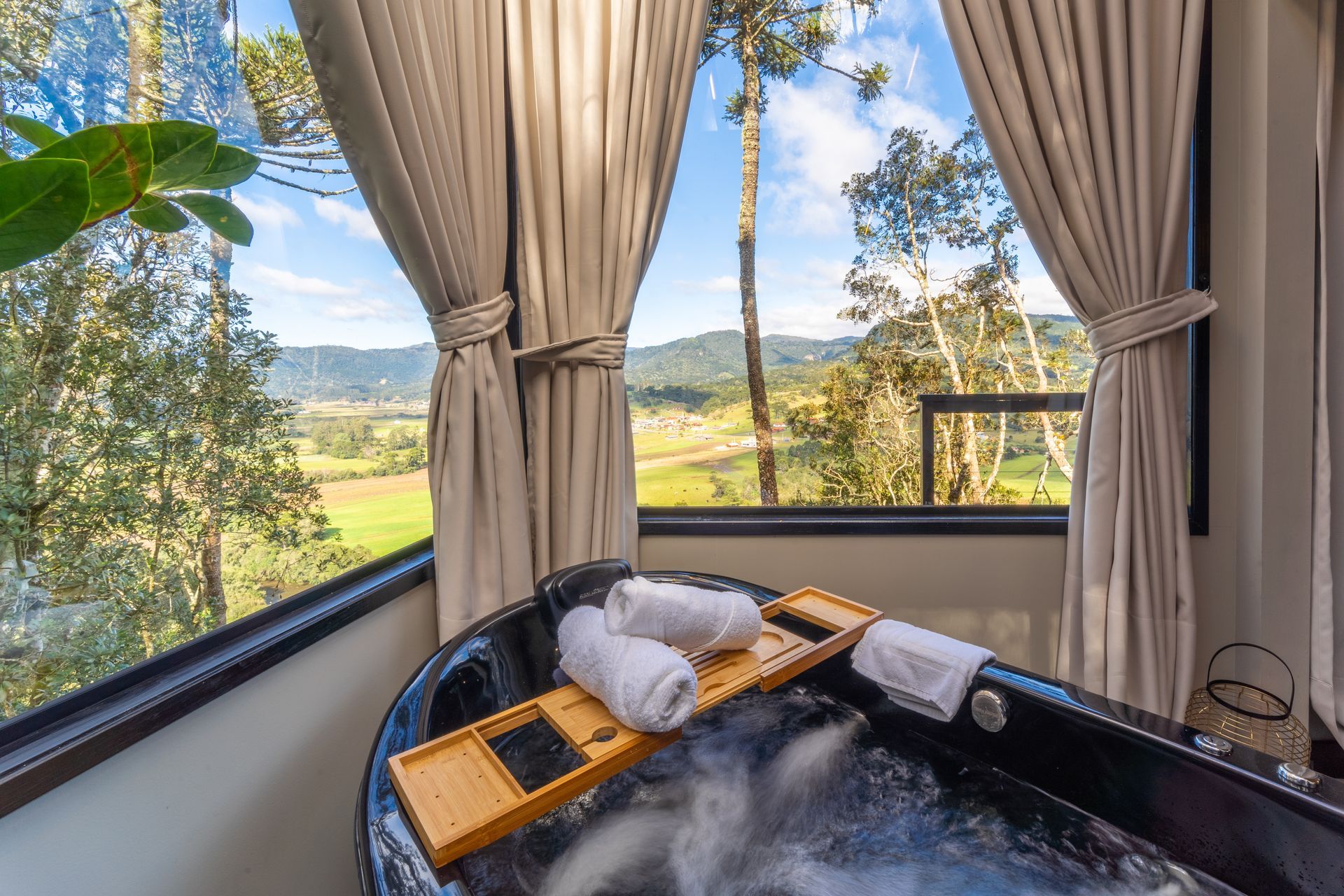 Banheira de hidromassagem preta com toalhas, bandeja de madeira e vista para a montanha através de uma grande janela.