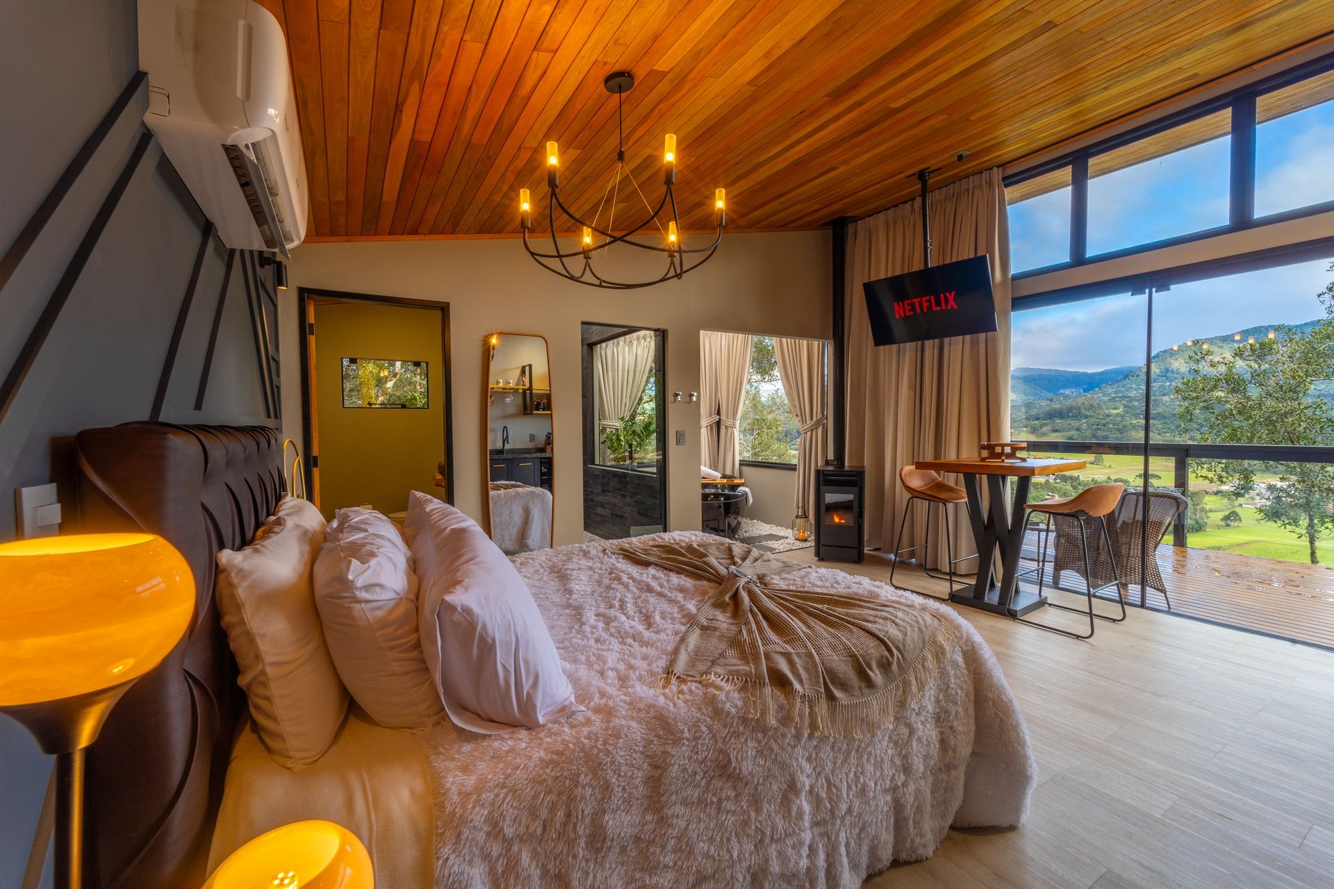 Quarto aconchegante com janela ampla e vista panorâmica. A cama possui edredom macio. Luminária de teto escura e abajures.