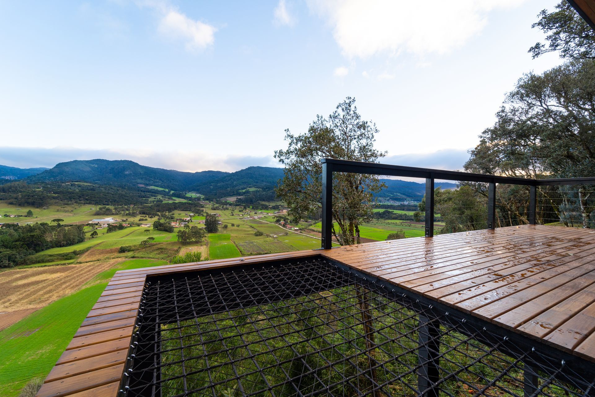 Varanda de madeira com corrimão de metal com vista para um vale, campos e montanhas em um dia ensolarado.
