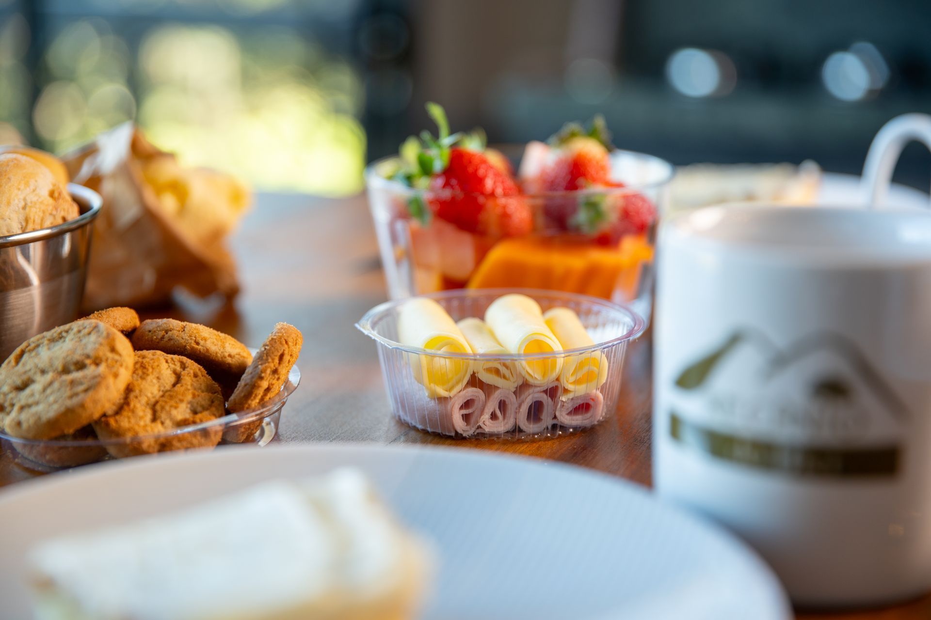 Café da manhã: salada de frutas, biscoitos, pãezinhos, pãezinhos de presunto e queijo, caneca branca e um prato.