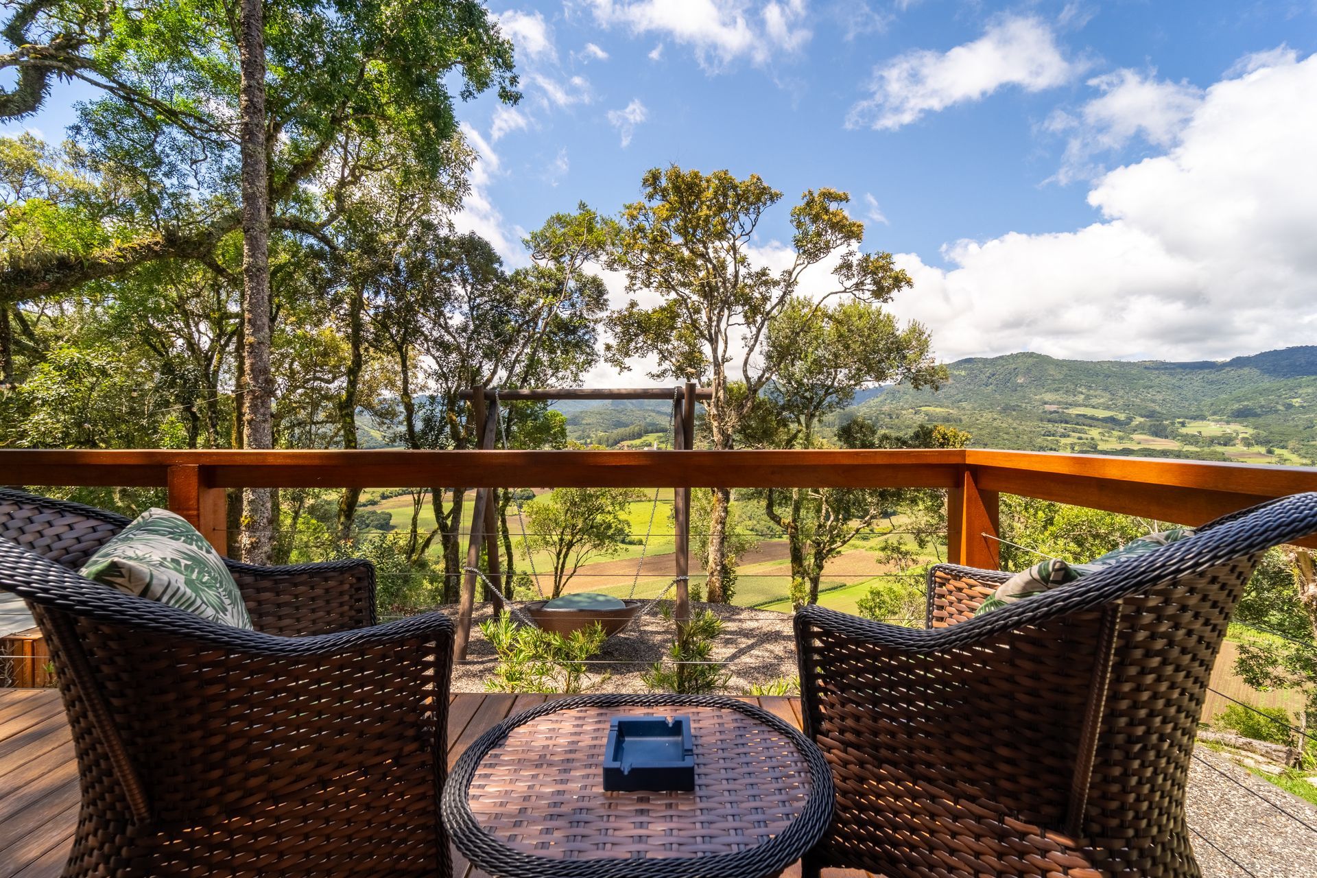 Duas cadeiras de vime e uma mesa em um deck com vista para um vale, árvores e montanhas em um dia ensolarado.