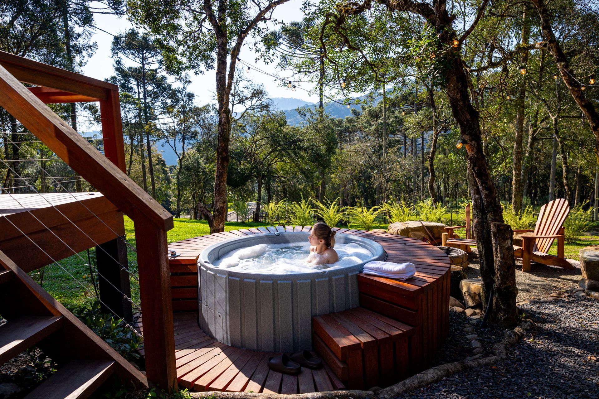 Mulher em uma banheira de hidromassagem cercada por um deck de madeira, árvores e vista para as montanhas.