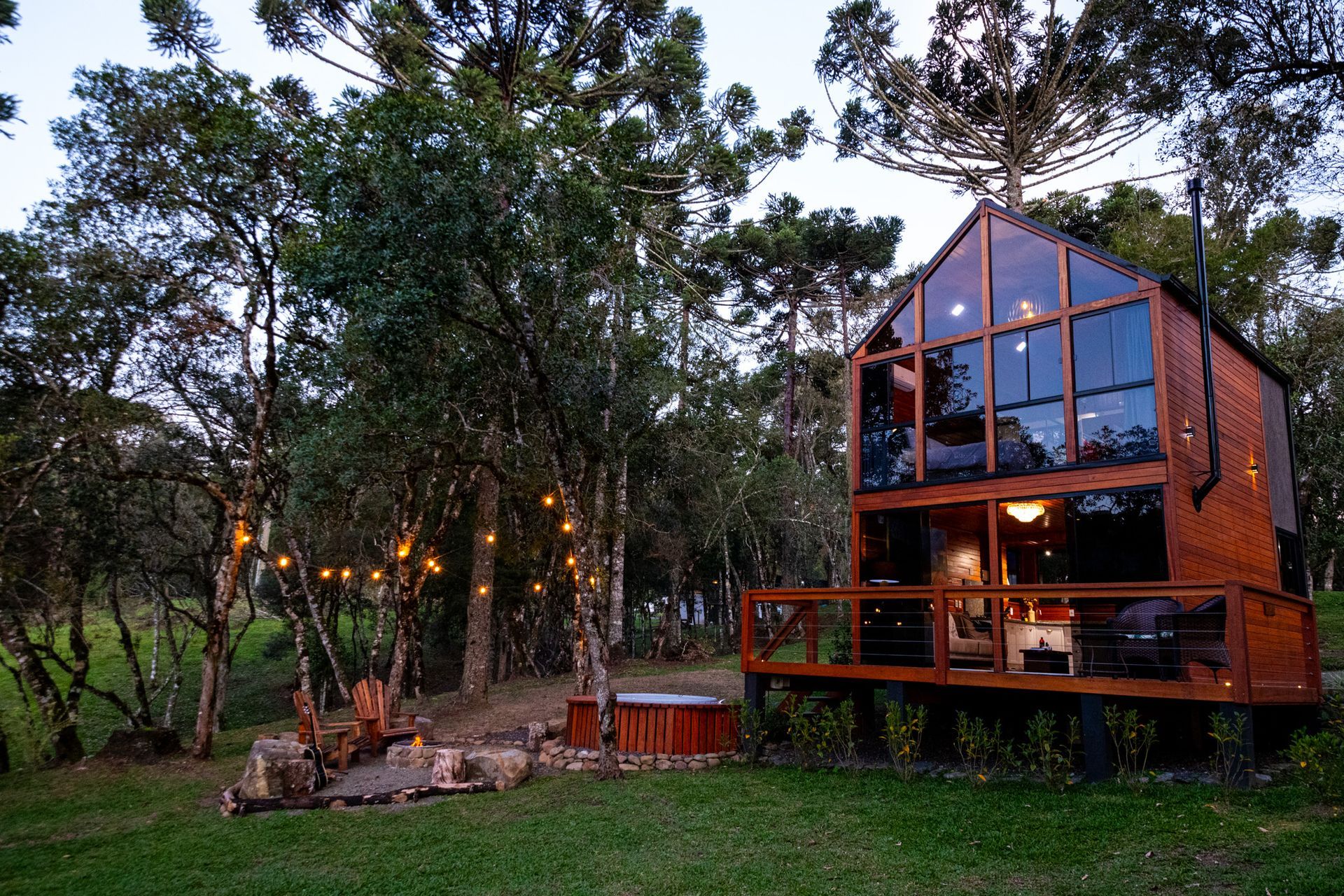 Cabana de madeira com grandes janelas, situada em uma área arborizada, cercada por árvores e uma fileira de luzes acesas.