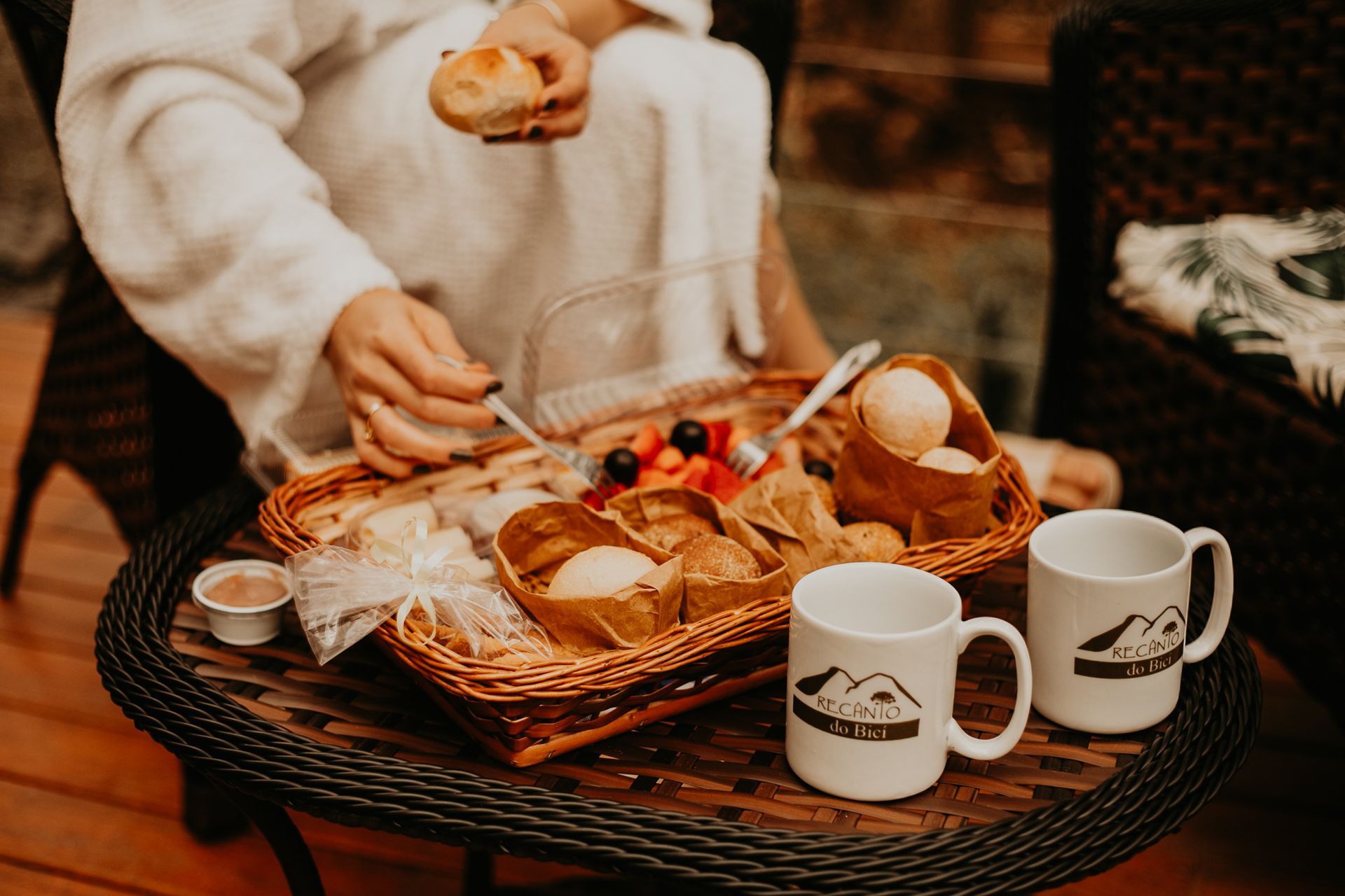 Pessoa de roupão desfruta de um café da manhã com frutas, doces e canecas sobre uma mesa de vime.