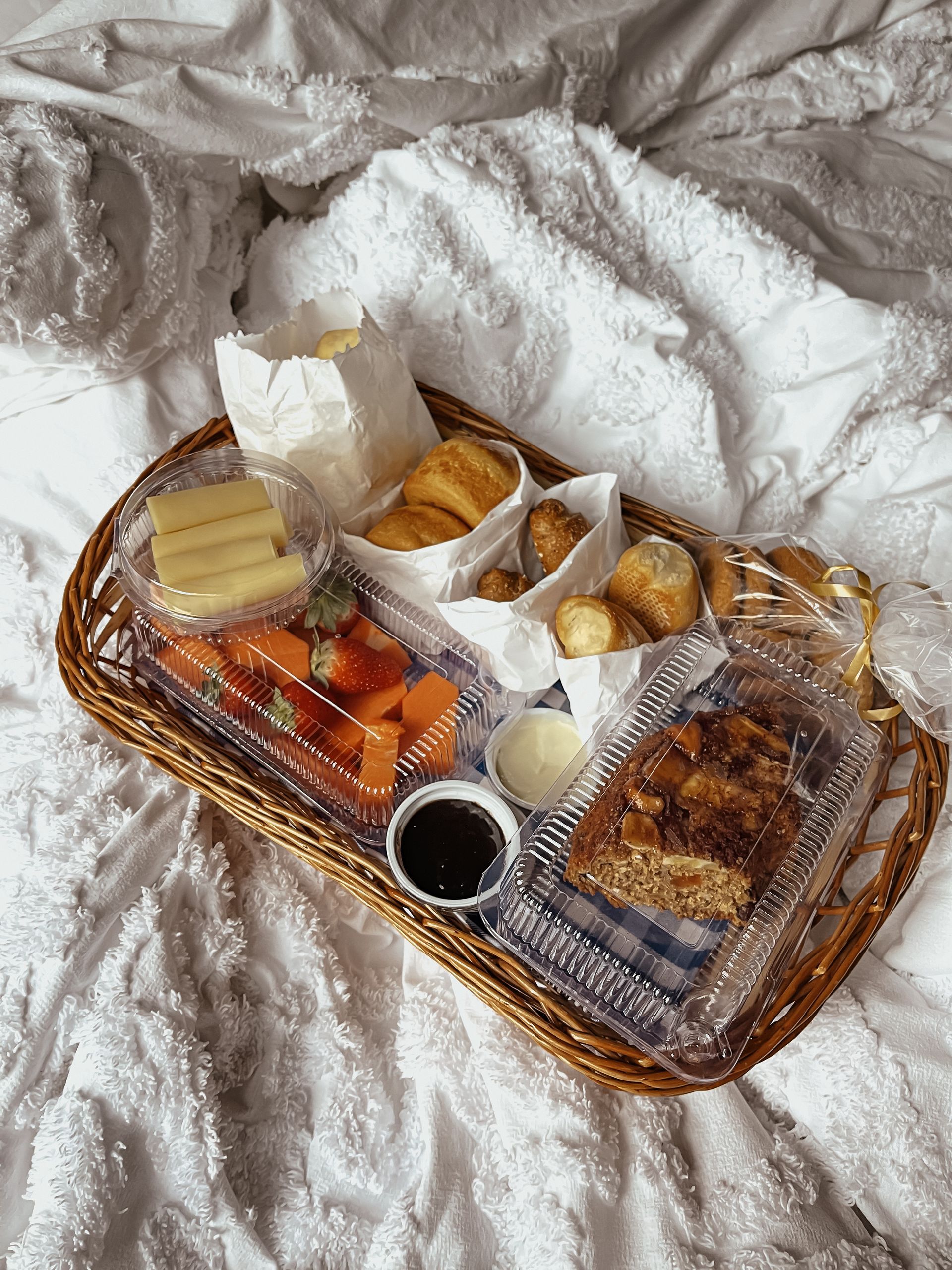 Bandeja de café da manhã sobre uma colcha branca texturizada. Contém frutas, doces, queijo e geleia.